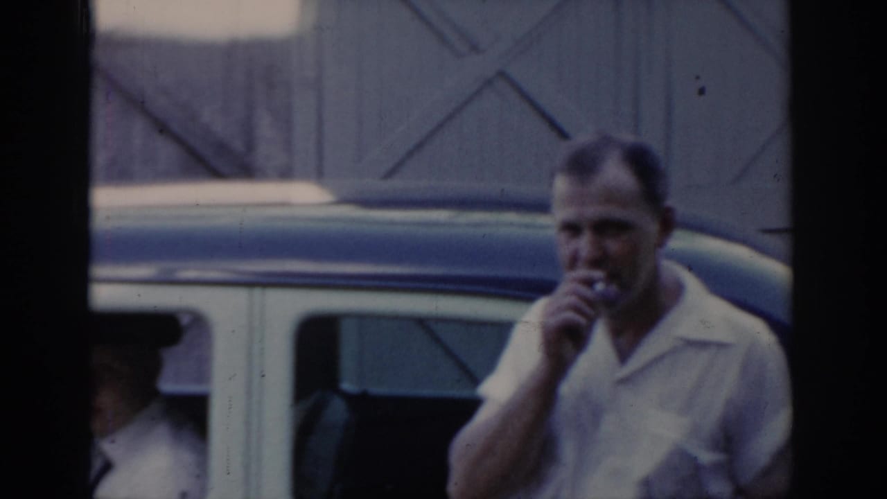 A man in a white short-sleeve shirt holds a cigarette near his mouth beside a white-and-blue sedan; a second figure is partially visible at left.