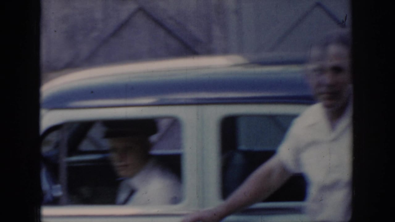 8mm frame of a smiling man in white shirt beside a white-and-blue taxi door, with a second capped figure visible through the car window, circa 1965 Saint Louis.