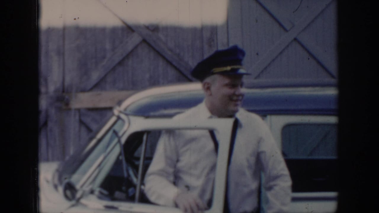 8mm frame of a taxi driver in white uniform jacket, dark tie, and peaked cap smiling outside his white-and-blue cab, Saint Louis, Missouri, circa 1965.