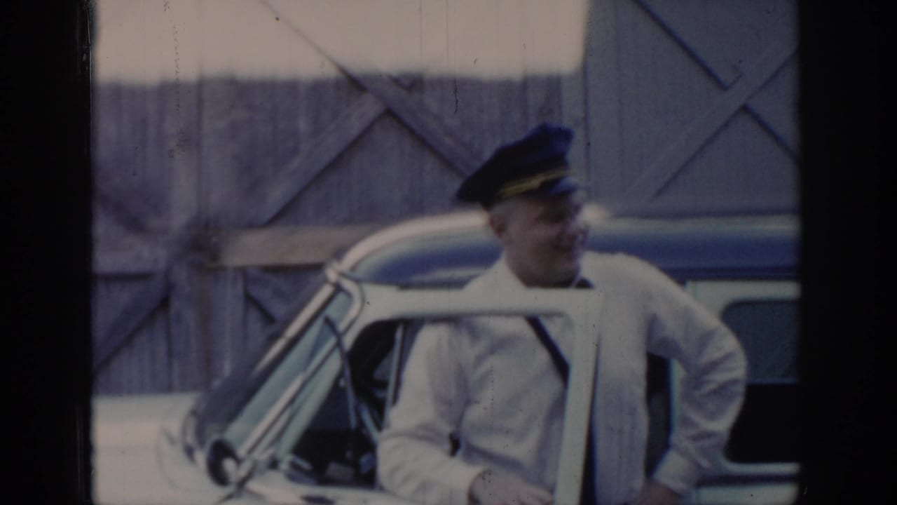 Man in dark peaked cap and white uniform shirt stands beside an open car door, X-patterned wooden barn wall behind him.
