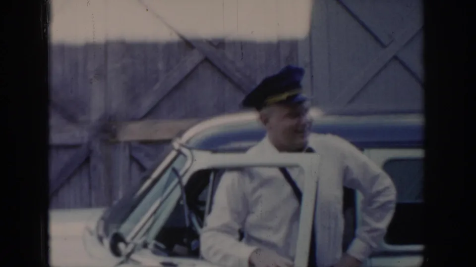 Man in a service worker's dark peaked cap and white long-sleeved shirt leans near an open car door, Saint Louis area, circa 1965, 8mm home movie.