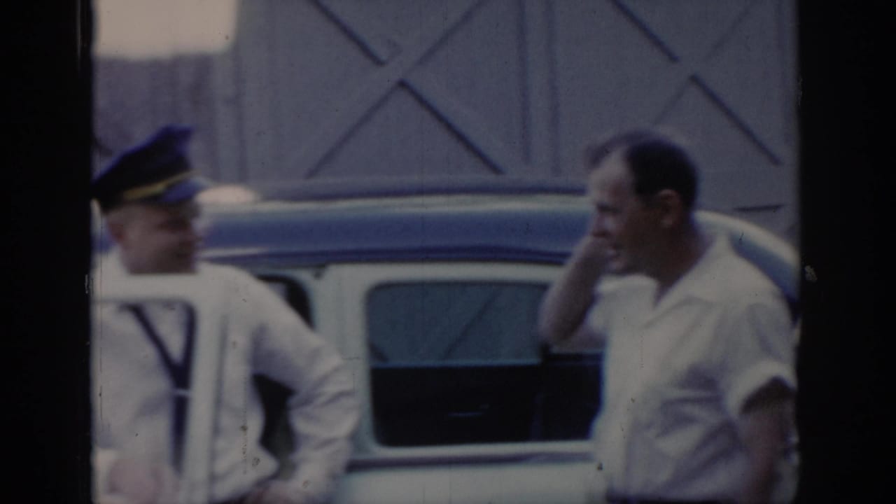 Two adult men conversing beside a light-colored two-tone car in front of an X-patterned wooden building, Saint Louis area, circa 1965, 8mm home movie.