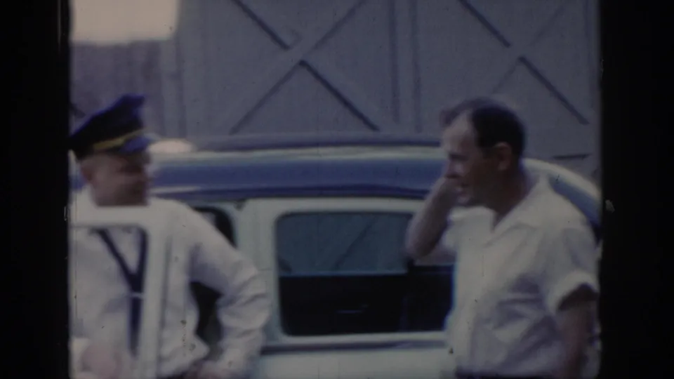 Two adult men conversing beside a light-colored two-tone car in front of an X-patterned wooden building, Saint Louis area, circa 1965, 8mm home movie.