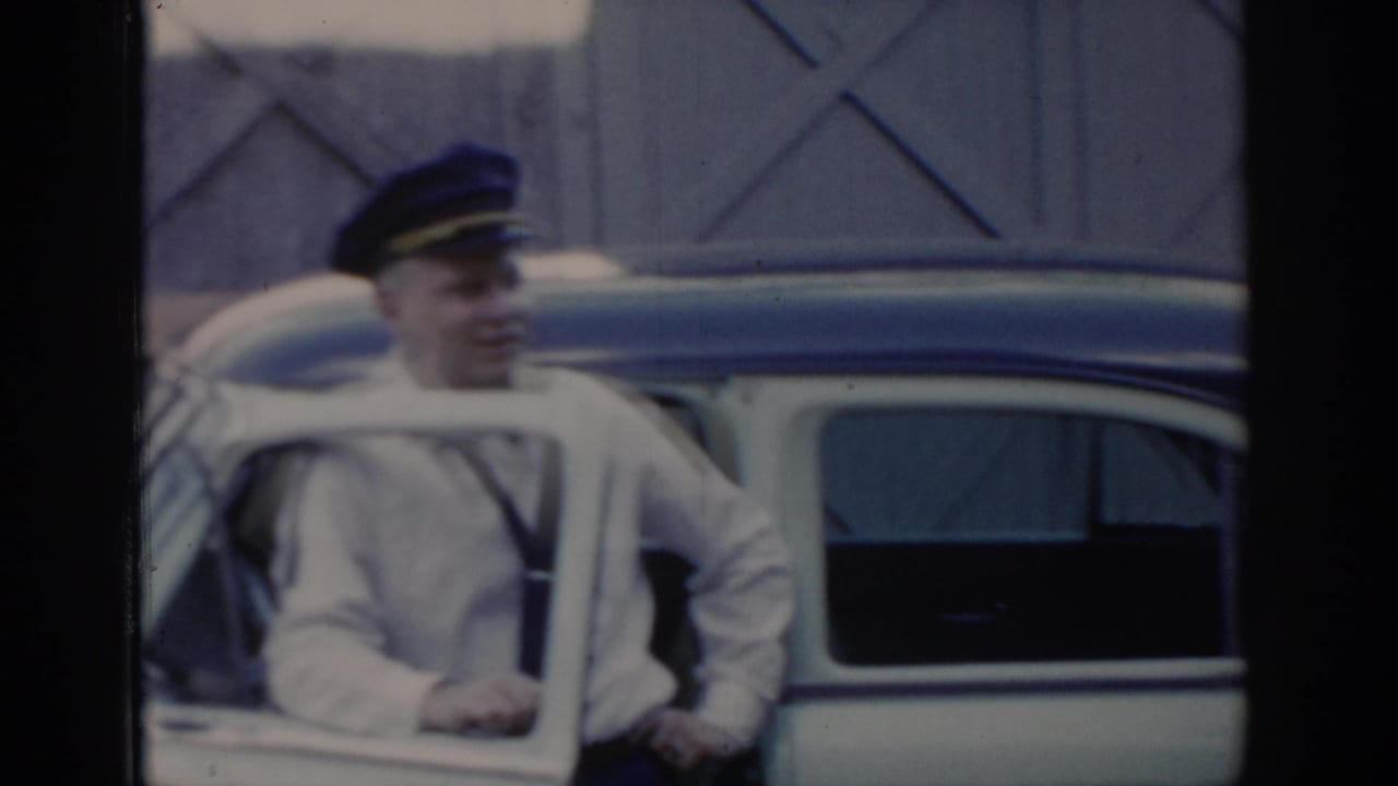 Smiling man in dark peaked cap and white uniform shirt stands near a car door, X-patterned barn-style building behind him, Saint Louis area, circa 1965.
