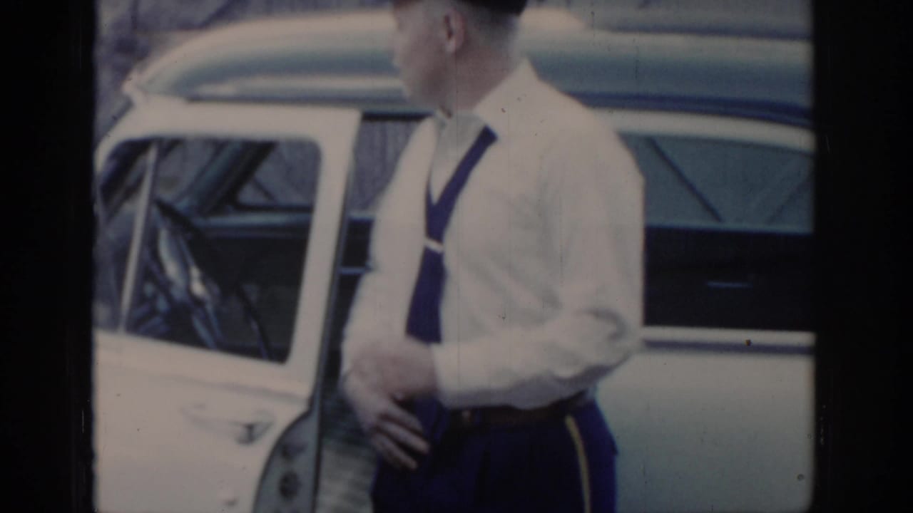 Man in white shirt with visible dark suspenders or uniform straps stands beside a two-tone American car, X-patterned wooden building in background, circa 1965.