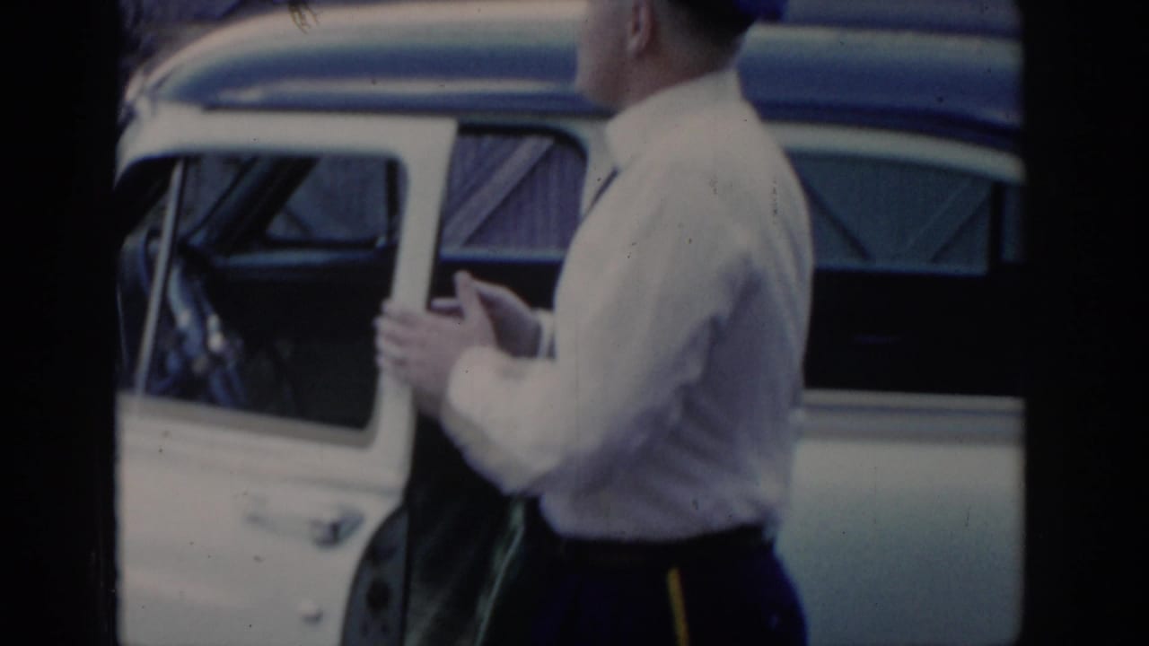 Mid-1960s uniformed attendant in white shirt stands beside an open car door; a wooden-clad structure is visible in the background, Saint Louis, Missouri, 1965.