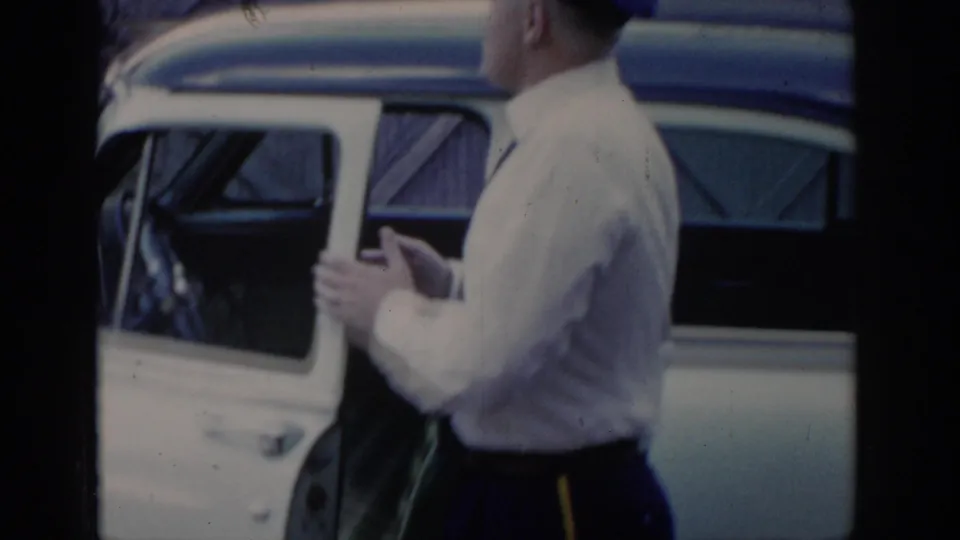 Mid-1960s uniformed attendant in white shirt stands beside an open car door; a wooden-clad structure is visible in the background, Saint Louis, Missouri, 1965.