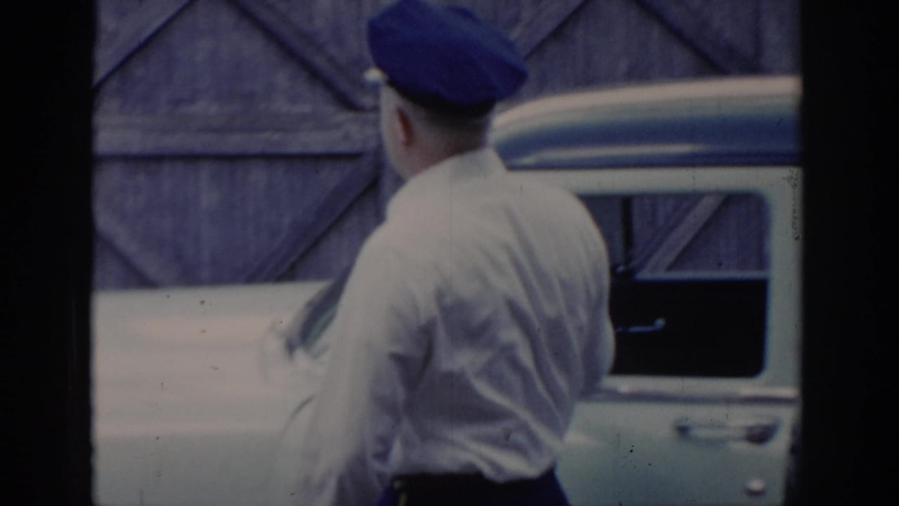 Man in blue cap and white shirt stands with his back to camera beside a light-colored car, a wooden-plank wall behind him.