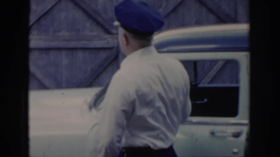 Uniformed man in blue cap stands with back to camera beside a parked car; weathered wooden boards form the backdrop in this archival 8mm shot from 1965.