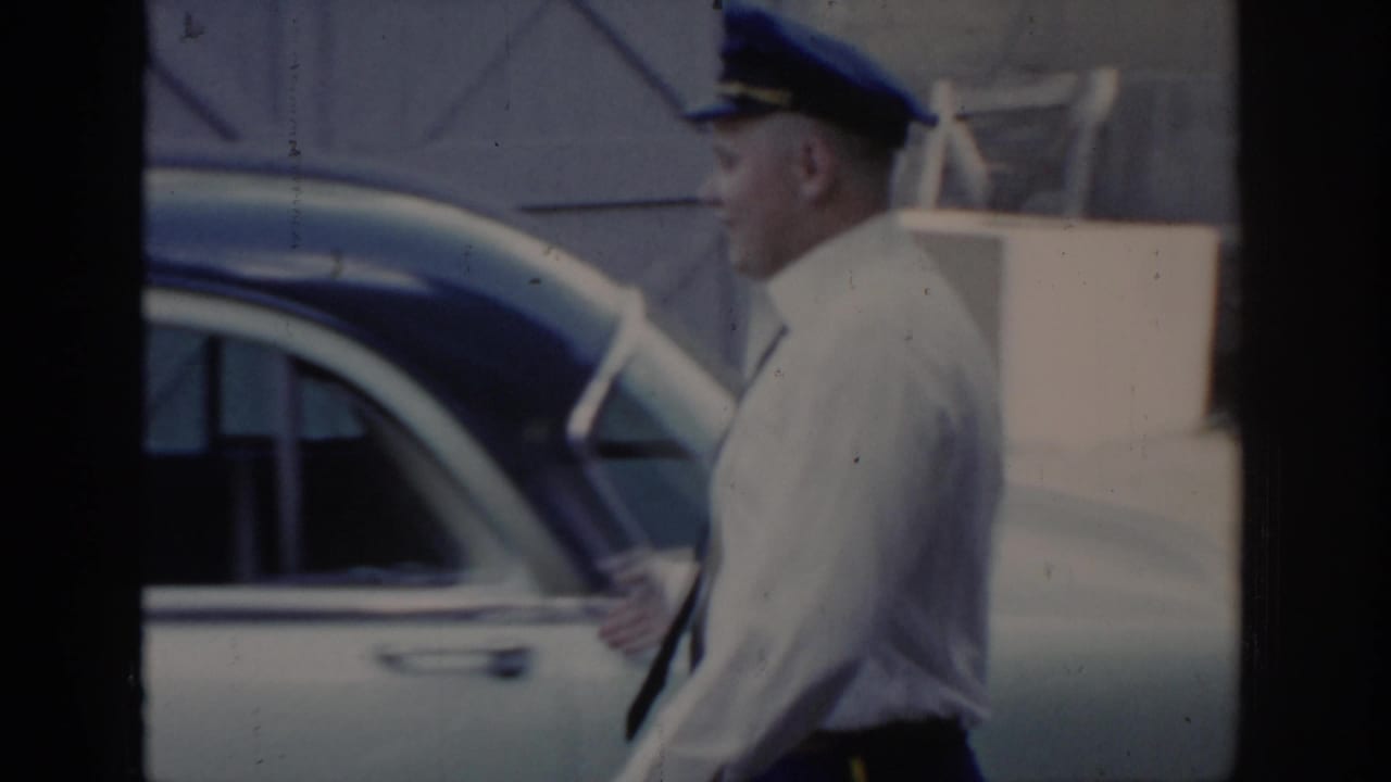 Uniformed attendant in blue cap reaches for a car door in what appears to be a service or arrival area; wooden structure and paved surface visible, Saint Louis, 1965.
