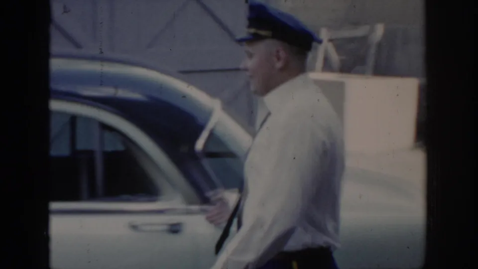 Uniformed attendant in blue cap reaches for a car door in what appears to be a service or arrival area; wooden structure and paved surface visible, Saint Louis, 1965.