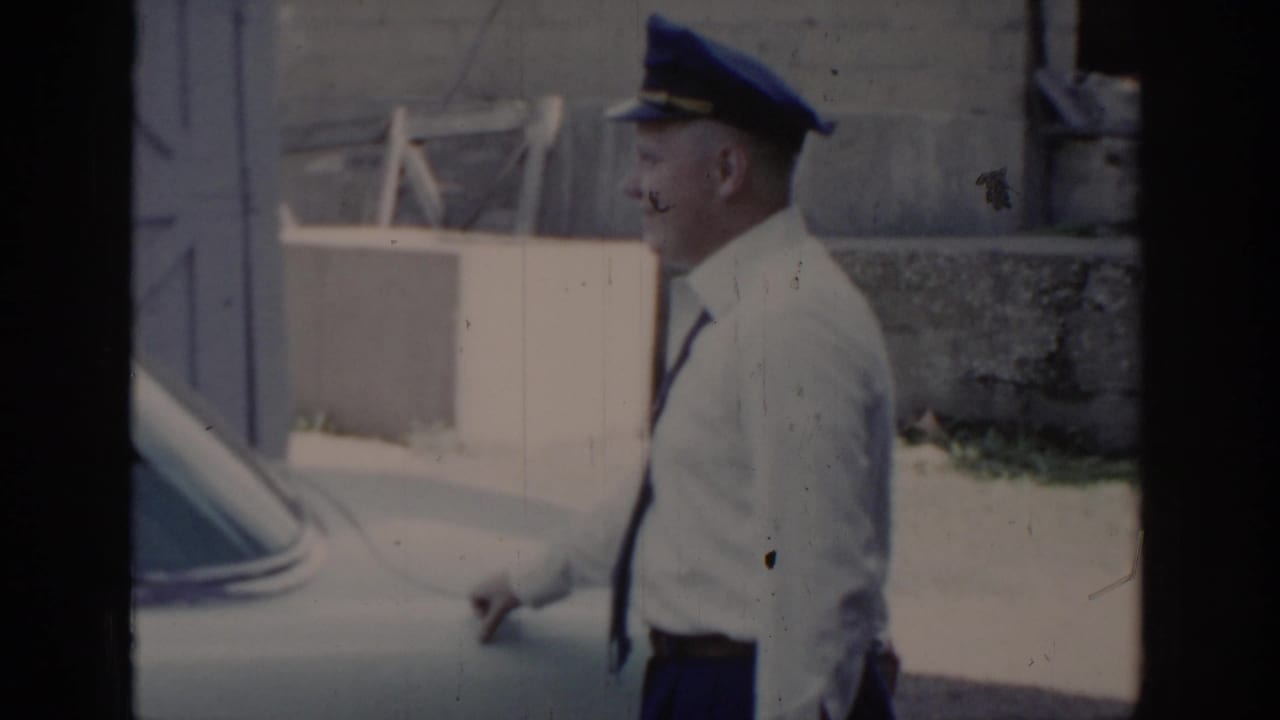 Older uniformed man in blue cap, white shirt, and dark tie stands in profile outdoors beside a light-colored car, with a concrete block wall in the background, Saint Louis, 1965.