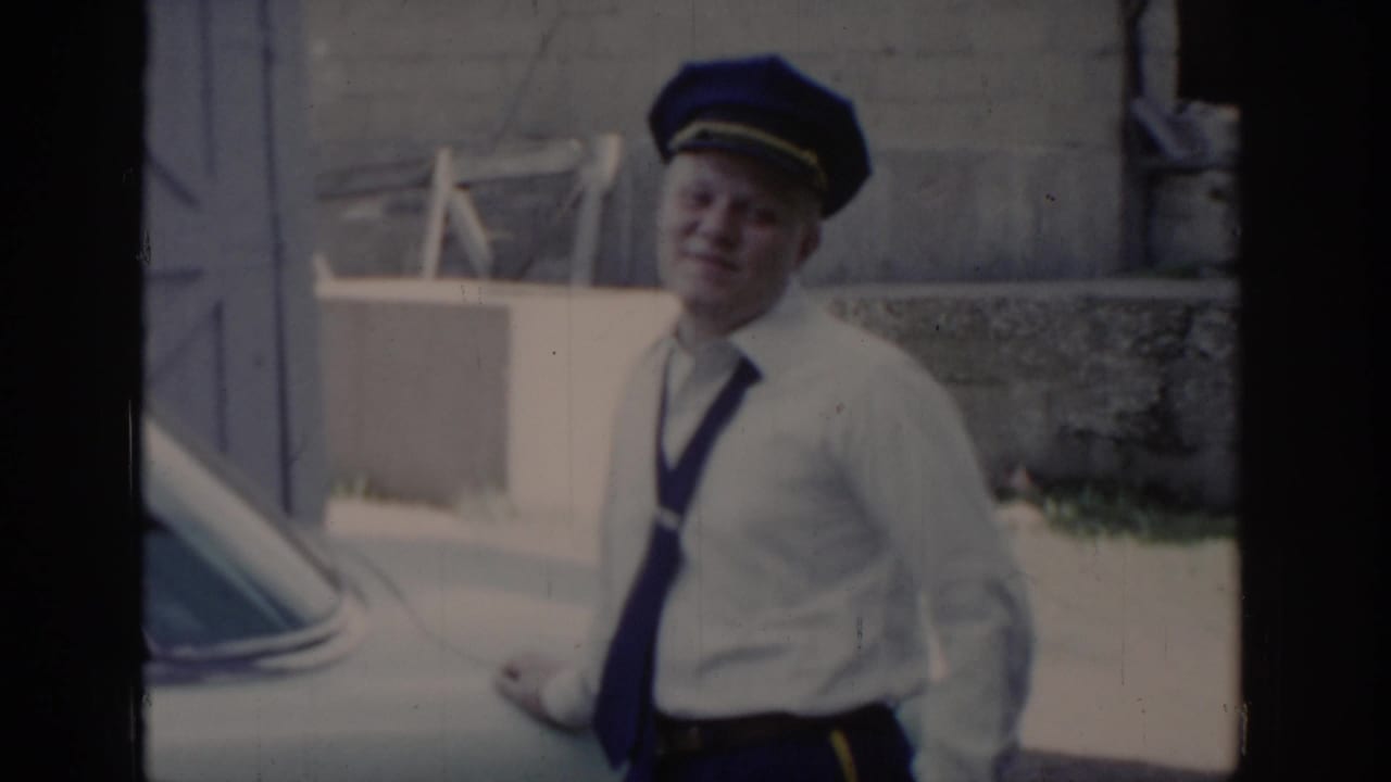 Uniformed officer in white shirt, dark tie, and blue peaked cap standing outdoors beside a parked vehicle, smiling, St. Louis, Missouri, circa 1965.