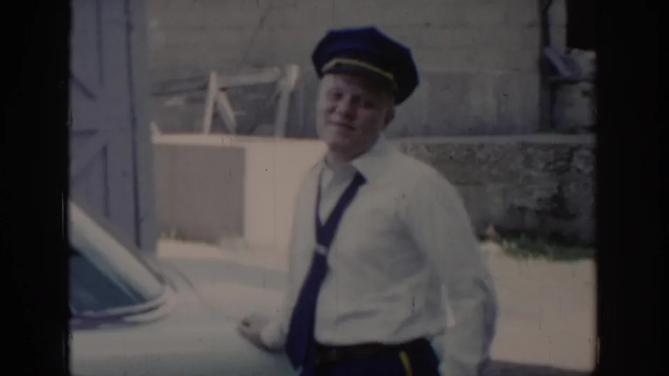 Uniformed officer in white shirt, dark tie, and blue peaked cap standing outdoors beside a parked vehicle, smiling, St. Louis, Missouri, circa 1965.