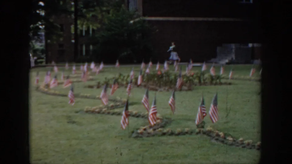 1965 Saint Louis area 8mm home movie: American flag display in curved garden rows, woman in light dress, brick building and trees in background.
