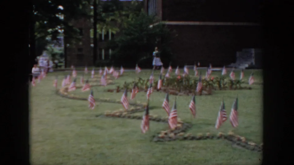 8mm home movie frame circa 1965, Saint Louis area: ornamental flag arrangement on garden beds, woman in background, brick building at right.