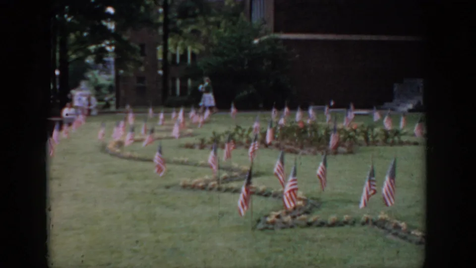 Mid-1960s 8mm frame, likely Saint Louis area: wide view of patriotic flag display, figure near brick building, dark doorway framing visible at left edge.