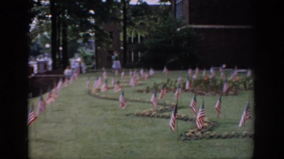 Final 8mm frame circa 1965, Saint Louis area: American flag garden display, figures in background near brick building, tree canopy above.