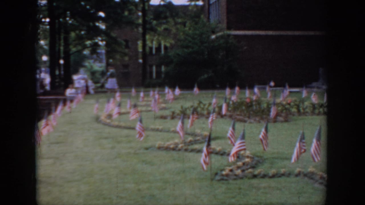 8mm home movie, circa 1965, Saint Louis, Missouri — American flags in curving rows on an institutional lawn; a small figure visible in the far background near the building.