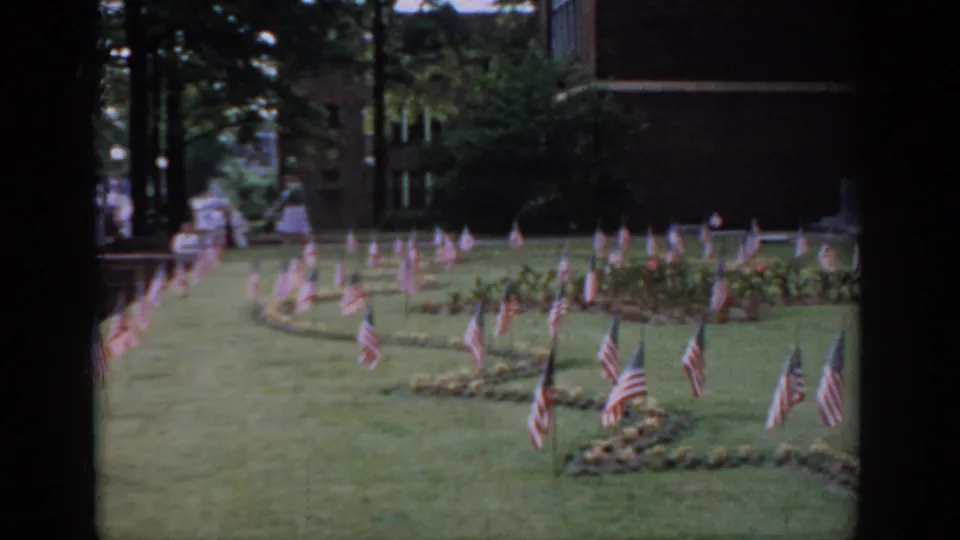 8mm home movie, circa 1965, Saint Louis, Missouri — American flags in curving rows on an institutional lawn; a small figure visible in the far background near the building.
