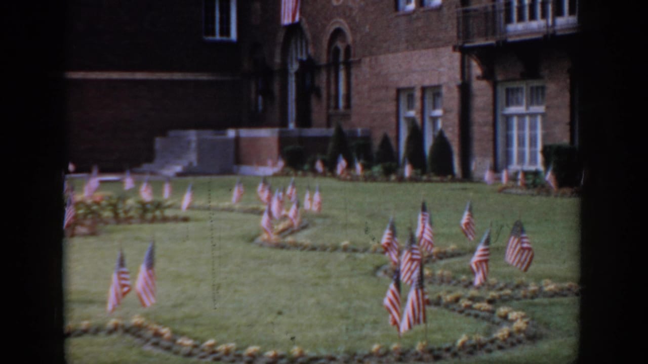 American flags in decorative pattern on grass, large red brick building with Romanesque arched windows and stone steps prominent at right, circa 1965.