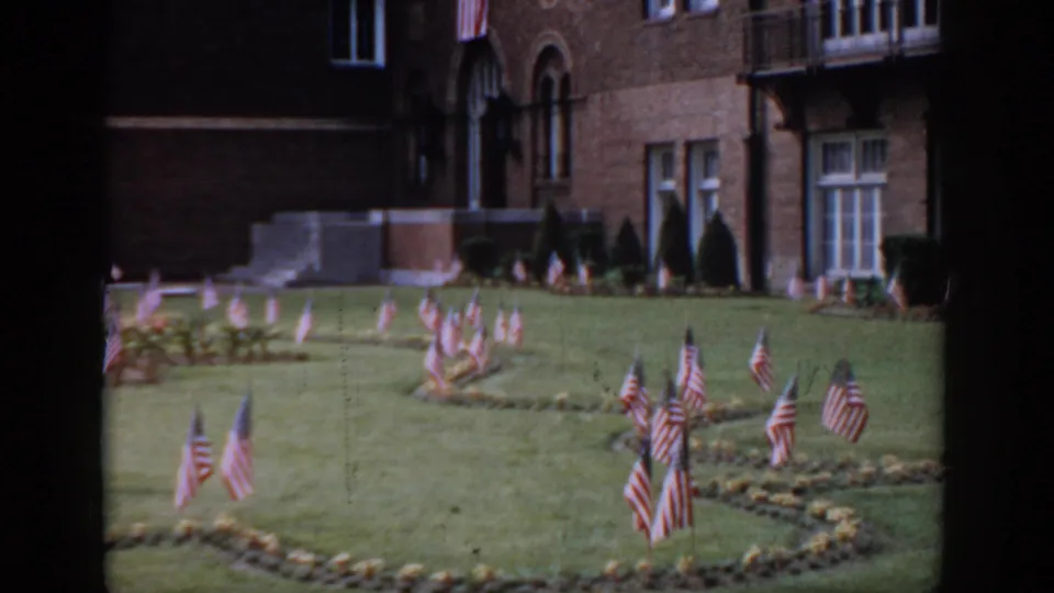 8mm footage, circa 1965, Saint Louis — American flag display on institutional grounds with a brick building featuring Romanesque arched windows, stone steps, and clipped topiary at right.
