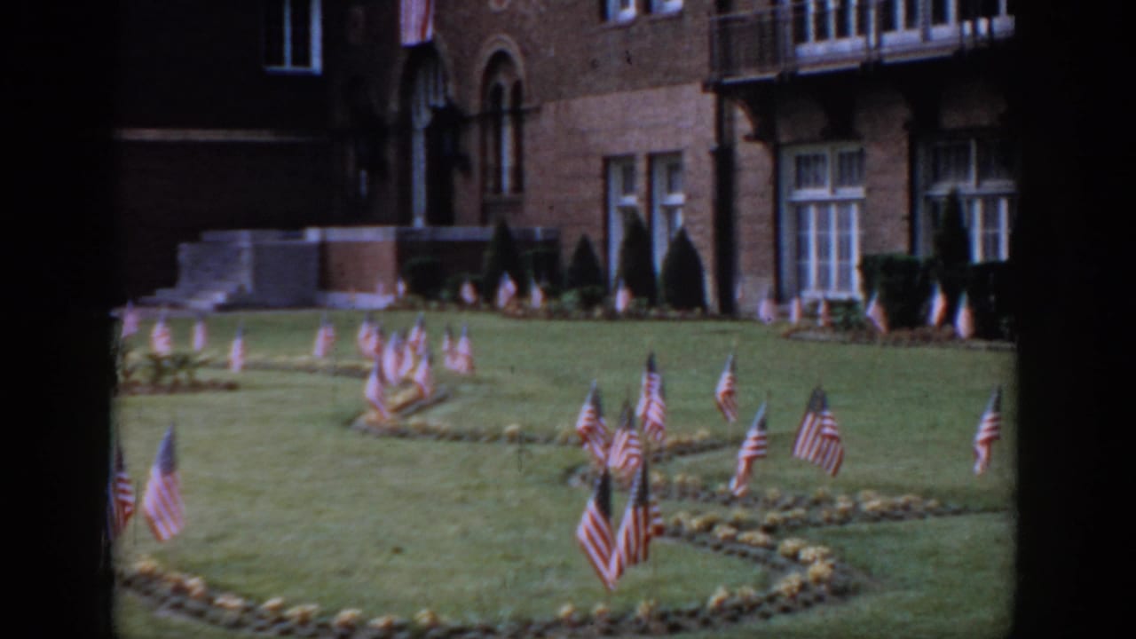 8mm, circa 1965, Saint Louis — red brick institutional building with Romanesque arched windows and stone entrance portico, clusters of American flags on the manicured lawn below.