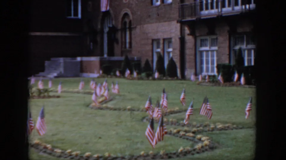 8mm, circa 1965, Saint Louis — red brick institutional building with Romanesque arched windows and stone entrance portico, clusters of American flags on the manicured lawn below.