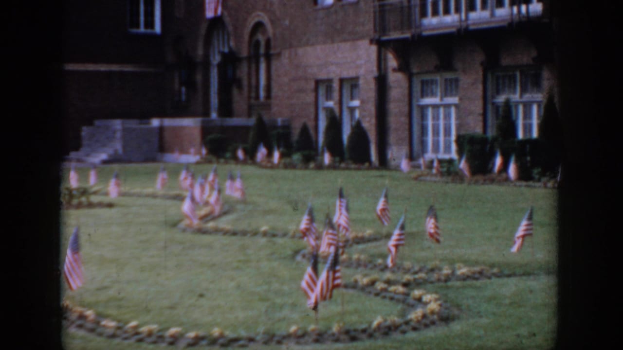 8mm, circa 1965, Saint Louis — decorative American flag clusters on manicured lawn, red brick building with arched windows and ornamental conical shrubs in background.