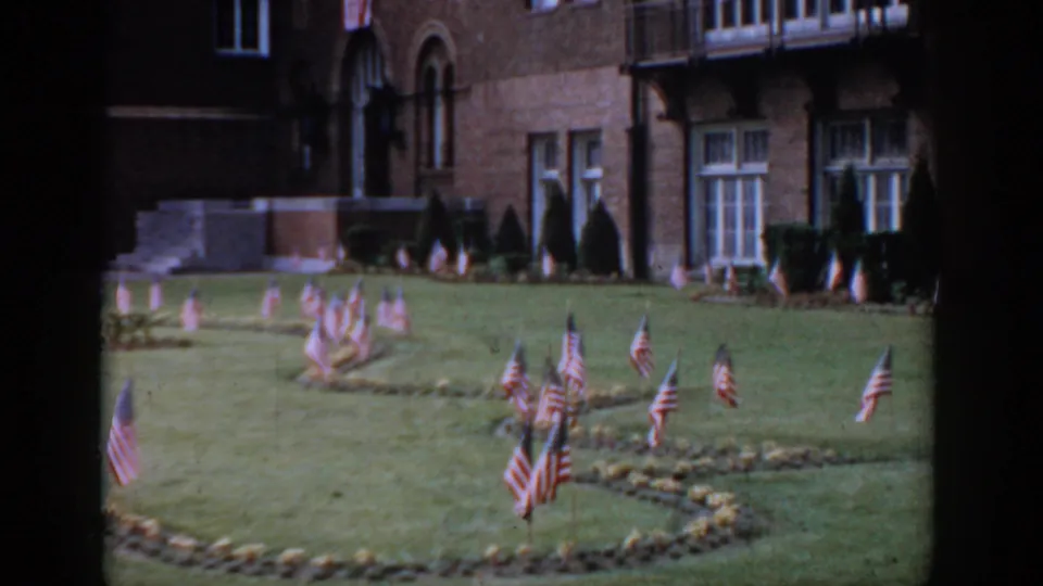 8mm, circa 1965, Saint Louis — decorative American flag clusters on manicured lawn, red brick building with arched windows and ornamental conical shrubs in background.