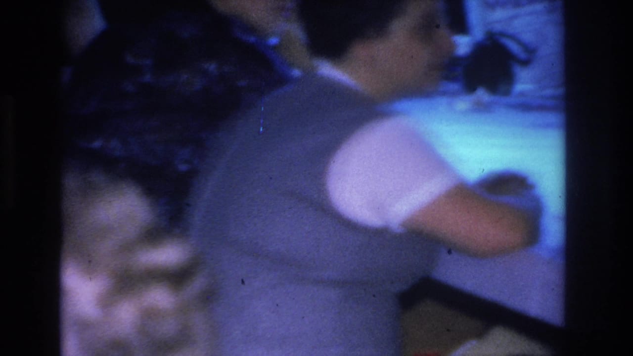 Woman in a light-colored short-sleeved top viewed from the side at an indoor social gathering, Hoboken, New Jersey, circa 1966, on color 8mm film.