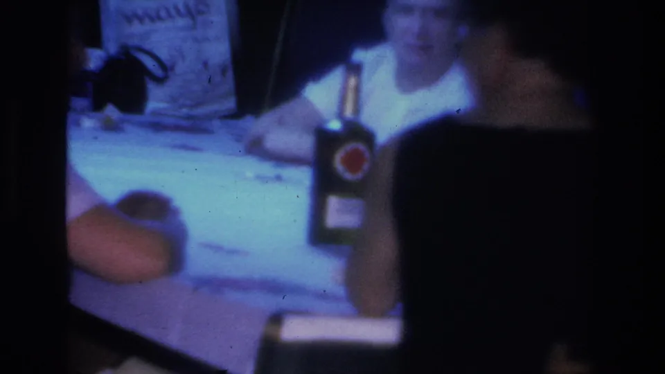 Women at an indoor gathering in Hoboken, New Jersey, circa 1966; a bottle with a red circular branded label and paper materials sit on the table, with partial background signage visible.