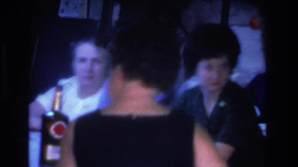 Two women in light-colored tops at an indoor social gathering in Hoboken, New Jersey, circa 1966; a bottle with a red circular branded label is prominent in the foreground.