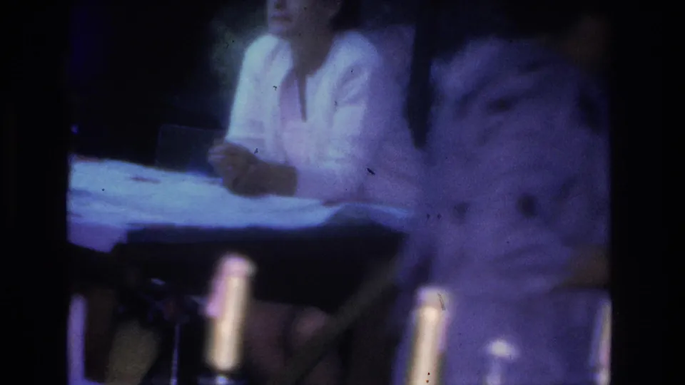 8mm home movie frame from a 1966 Hoboken party showing a woman in a light top and a second figure leaning over a table with bottles in blue-shifted low light.