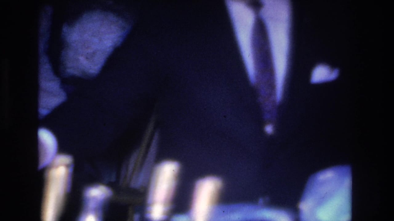 Man in dark suit with necktie and white pocket square bending toward a table with bottles in a dark blue-purple interior.