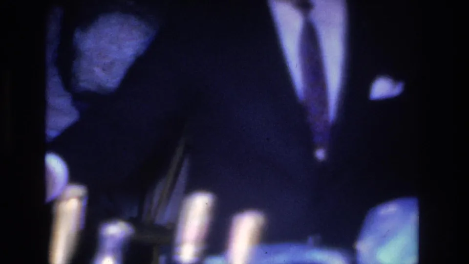 8mm frame from a 1966 Hoboken party showing a suited man with pocket square leaning toward a table bearing bottles, dark background with pronounced blue shift.