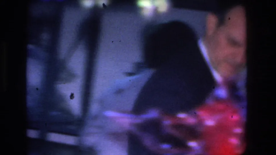 8mm frame from a 1966 Hoboken party showing a man in a suit jacket bending toward a table while handling a red-toned object, second figure partially visible, heavy grain and motion blur.
