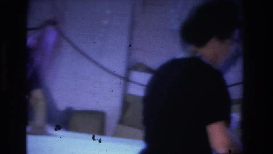 8mm home movie frame from a 1966 Hoboken party showing a figure in dark clothing leaning toward a table in a dim purple-blue interior with a chair in the background.