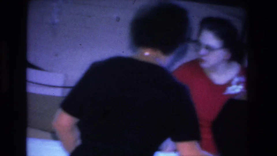 Two women at an informal indoor gathering, Hoboken, New Jersey, 1966; the woman facing camera wears a red knit top and eyeglasses; heavy purple 8mm color shift.
