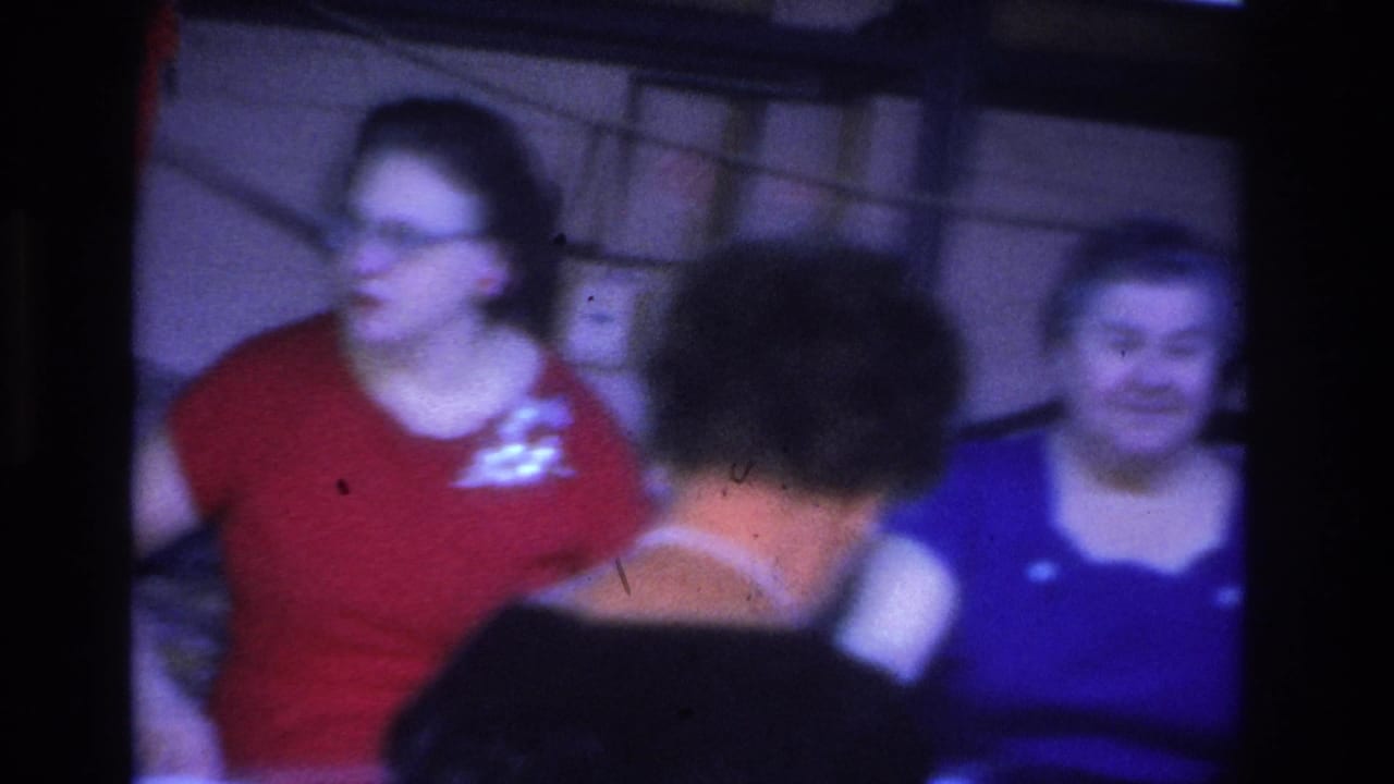 Woman in red top with glasses and a second woman in a blue blouse in a dimly lit interior; boxes or shelving visible in the background.