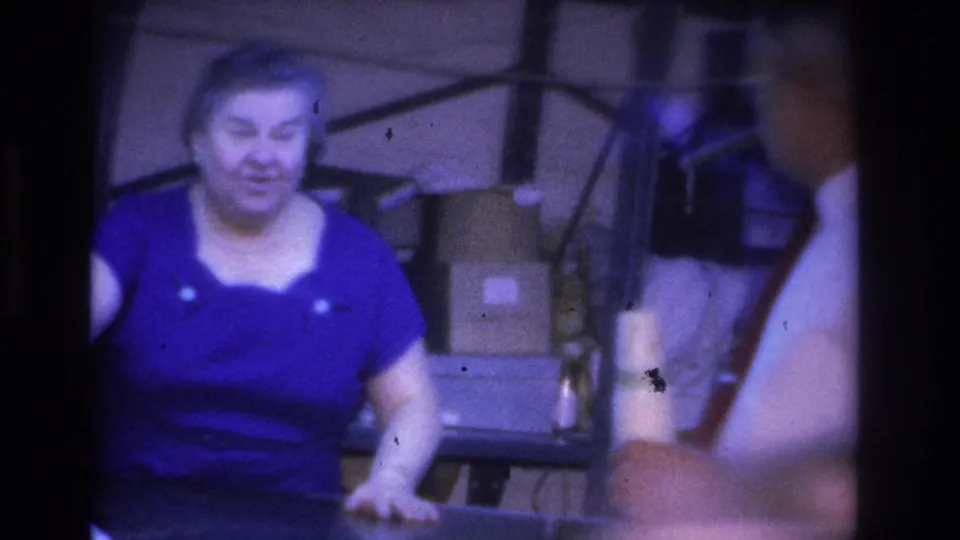 Older woman in a blue blouse in a back-room gathering space, Hoboken, New Jersey, 1966; cardboard boxes and a metal folding chair visible behind her; 8mm purple-blue color shift.