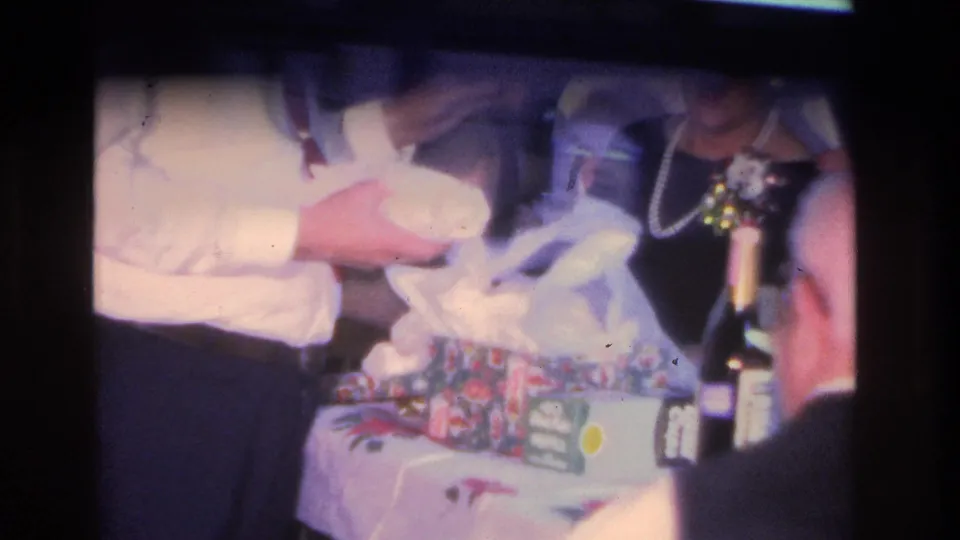 1966 Hoboken home movie: woman in dark dress and beaded necklace examining a wrapped gift at a floral-covered table, with a dark bottle and gathered guests visible.