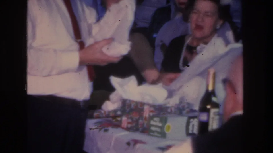 1966 Hoboken home movie: man in white shirt with suspenders and a woman wearing glasses at a floral table with gift wrapping and a dark bottle visible in the foreground.
