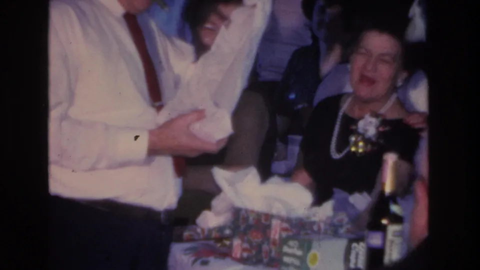 Indoor gift-opening scene, c. 1966, Hoboken NJ: man in white shirt and red tie at a table with wrapped presents; woman in black dress and pearls laughing.