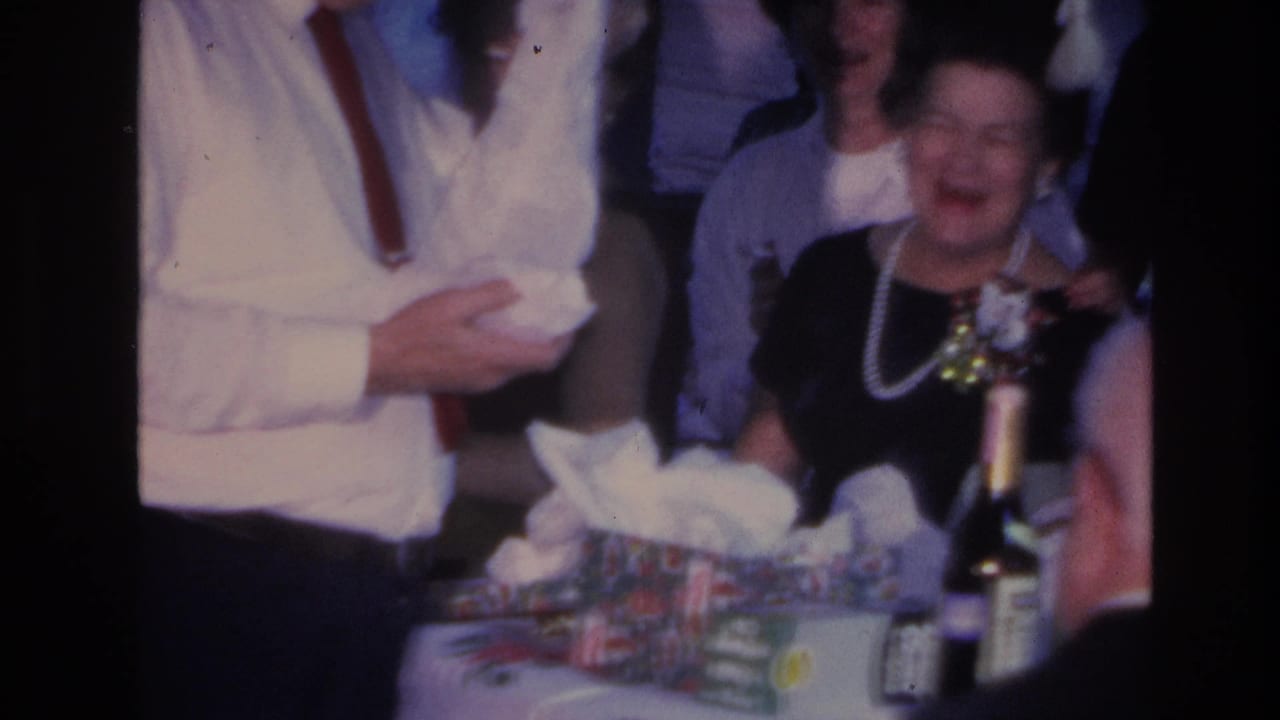 Man in white shirt and red tie still handling tissue paper from a gift; woman in black dress and pearl necklace smiles; gift boxes and a dark bottle on the table.