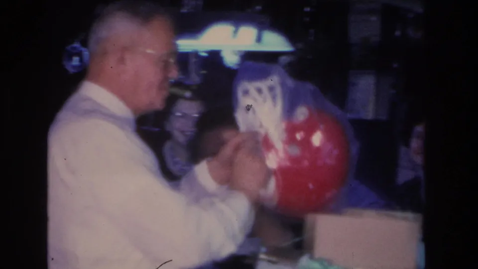 Older man with glasses and gray hair examines a red object consistent with a football helmet per clip title, at an indoor party, c. 1966, Hoboken NJ; motion blur from handheld camera.