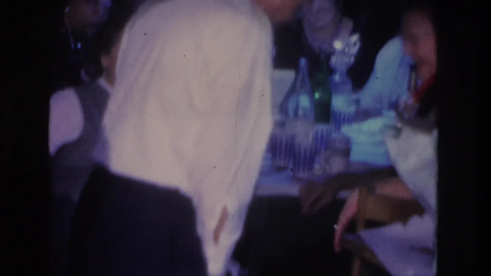 Wider party view, c. 1966, Hoboken NJ: woman in light blue dress at a table with glass bottles; man's white-shirted back in foreground; dark-haired guest visible at far right.