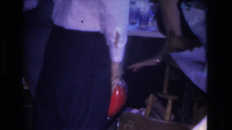 Torso-level 8mm frame of a man in mid-1960s dress attire holding a red spherical object in a dimly lit indoor party setting, Hoboken, New Jersey, 1966.