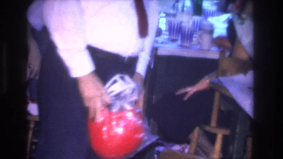 Mid-1960s 8mm frame of a man in white dress shirt gripping a red cellophane-wrapped sphere, with party glassware and wooden chairs visible behind him, 1966 Hoboken interior.