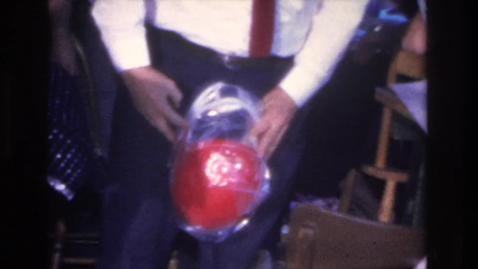 Close 8mm frame of a white-shirted man's hands holding a plastic-wrapped red sphere toward the camera in a 1960s indoor party setting, Hoboken, New Jersey.